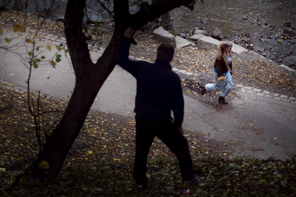 Mørkkledd mann bak et tre ser en ung jente gå forbi på veien. Foto.