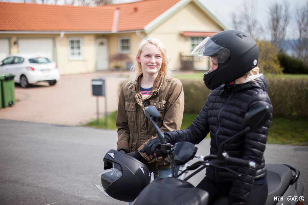 Jente på moped snakker med ei annen jente som holder en mopedhjelm i hendene. Foto.