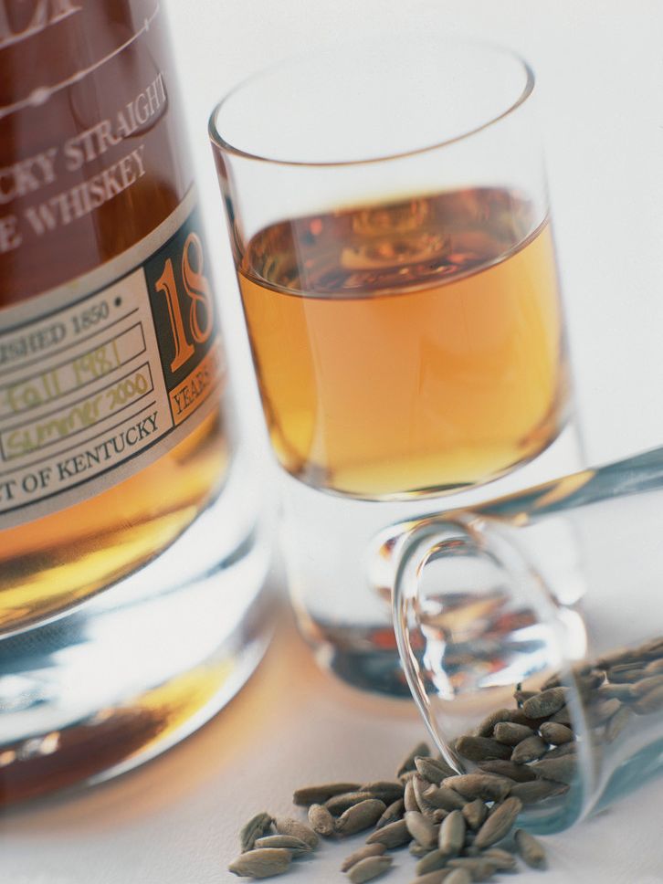 En flaske og et glass med rugwhiskey. Ved siden ligger et veltet glass med hel rug oppi. Foto.