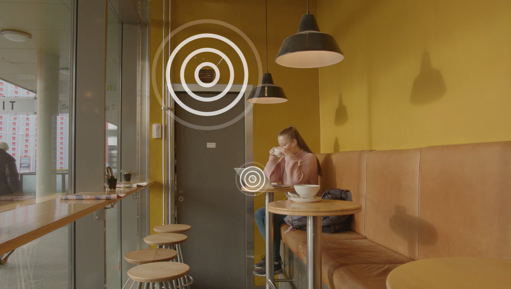 Jente sit ved eit bord på ein kafé. På biletet er det lagt inn grafiske symbol som viser elektromagnetiske bølger eller trådlause signal i lufta. Foto.