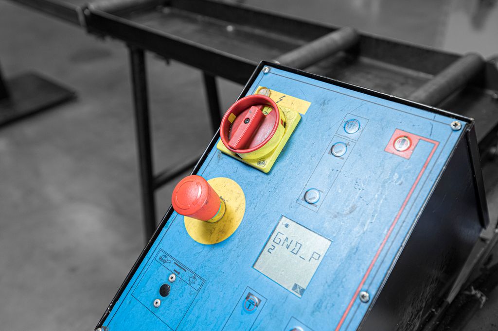 Ein stor, raud naudstoppbrytar i ein gul sirkel på eit blått beteningspanel. Foto.