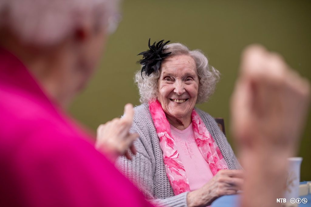 Eldre dame som smiler. Hun har rosa skjerf, grå jakke og en stor, svart blomst i håret. I forgrunnen ser vi en annen eldre kvinne med rosa klær, hun peker på dama som smiler. Foto.