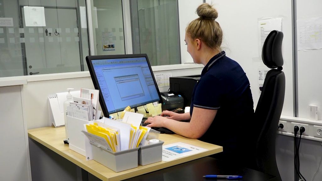 En kvinne sitter og jobber på en datamaskin i resepsjonen på et legekontor. Foto.