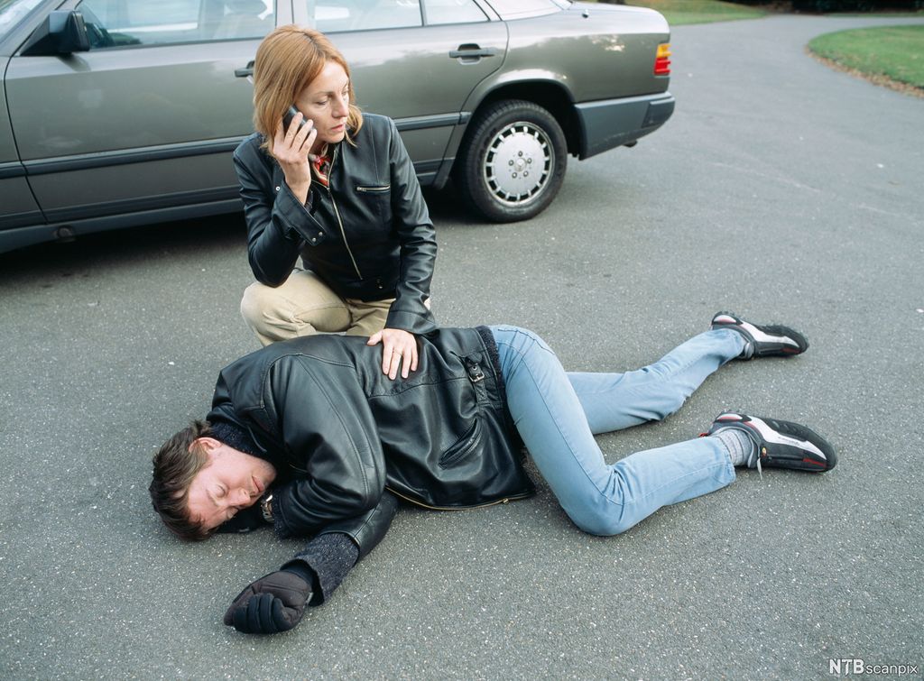 En mann er lagt i sideleie på asfalten foran en bil. En kvinne støtter ham og snakker i telefonen. Foto.