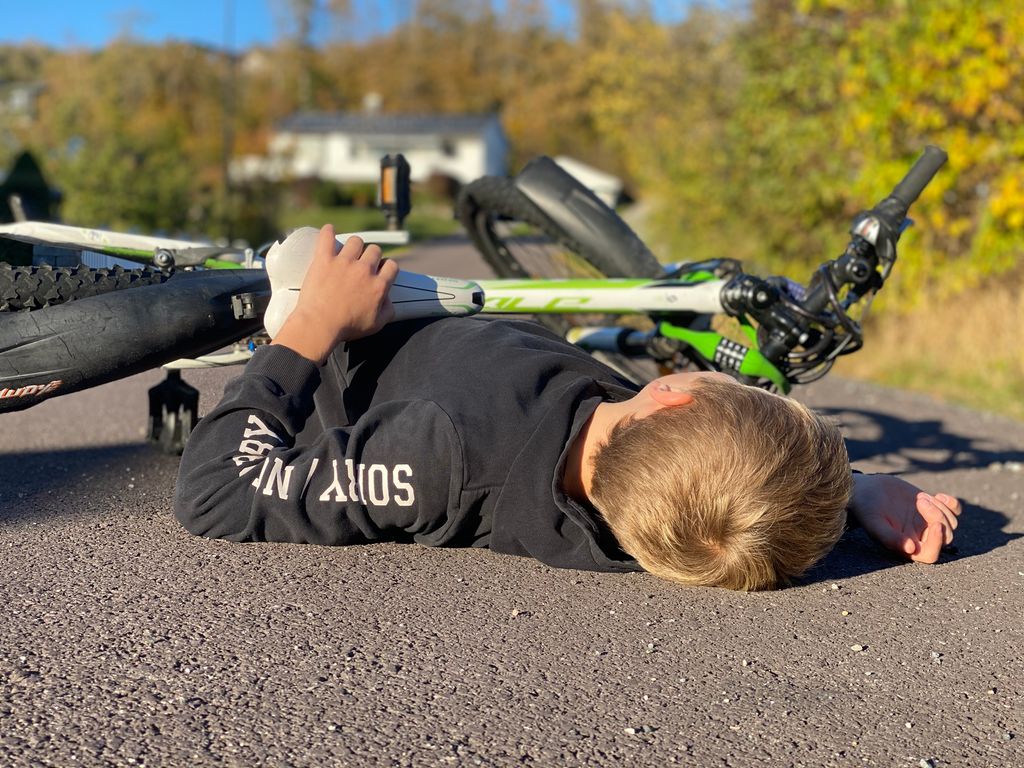 En ung gutt uten hjelm ligger på asfalten med en sykkel over seg. Foto. 