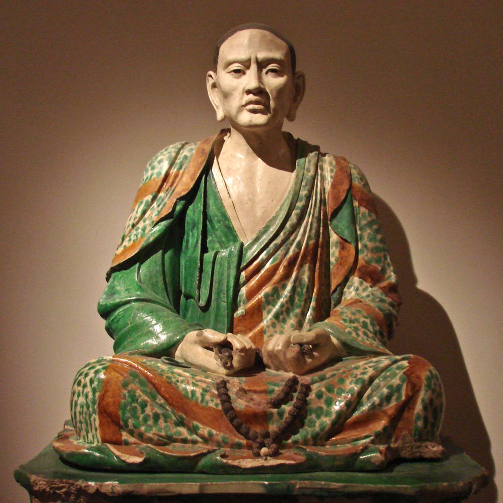 Mann sitter i lotusstilling iført grønn og rød kappe. Holder et kjede i hendene. Skulptur.