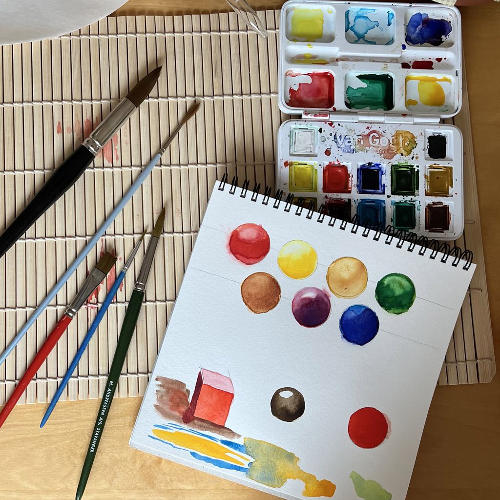 Pensler, akvarellskrin og akvarellpapir på et arbeidsbord. Kuler er malt i ulike farger og med høylys og skygger som gir tredimensjonal effekt. En kube er malt omgitt av fargeutprøvinger. Foto.