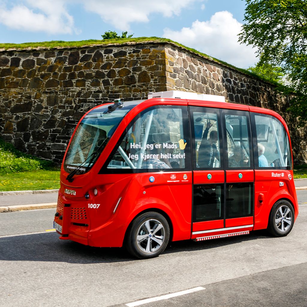 Liten, rød buss med passasjerer kjører på veien. På siden av bussen står det "Hei, jeg er Mads. Jeg kjører helt selv!". Foto.