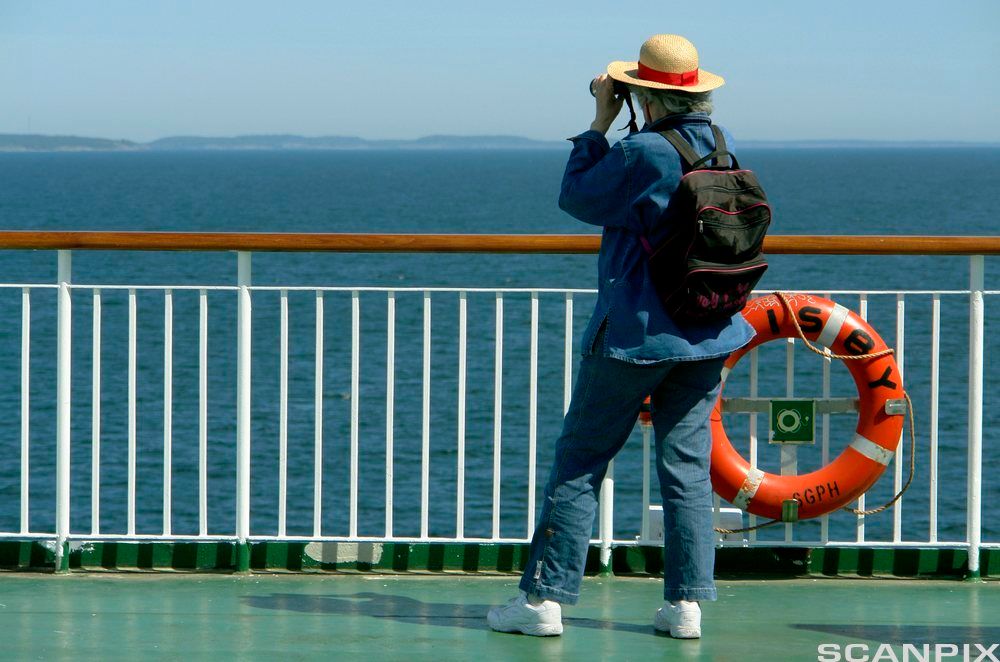 Person står på dekk, holder en kikkert foran øynene og kikker mot land. Foto.