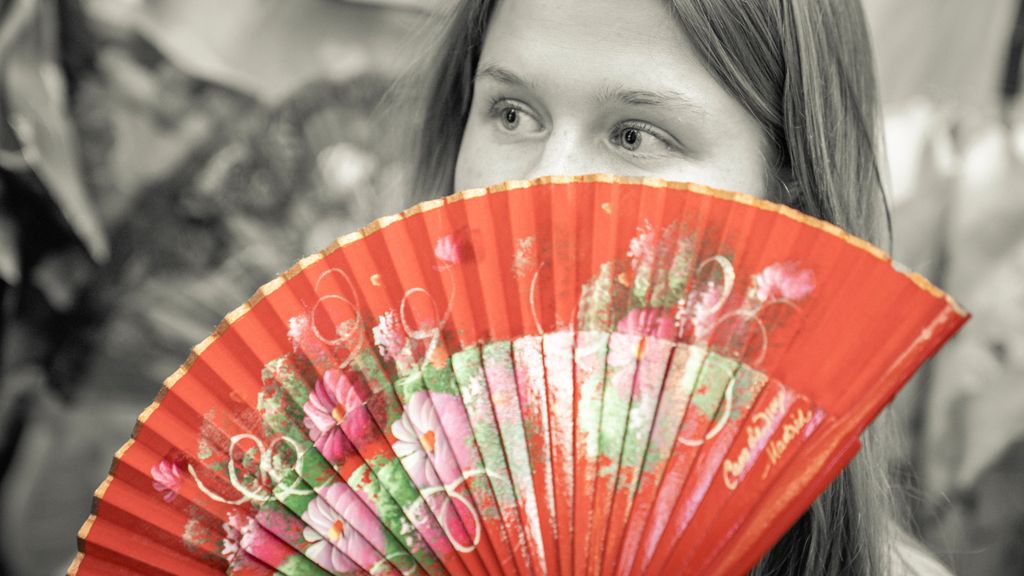 Ei håndvifte i røde farger med blomsterdekor foran ei jente i svart-hvitt. Munnen er dekket, men øynene er synlige. Foto.