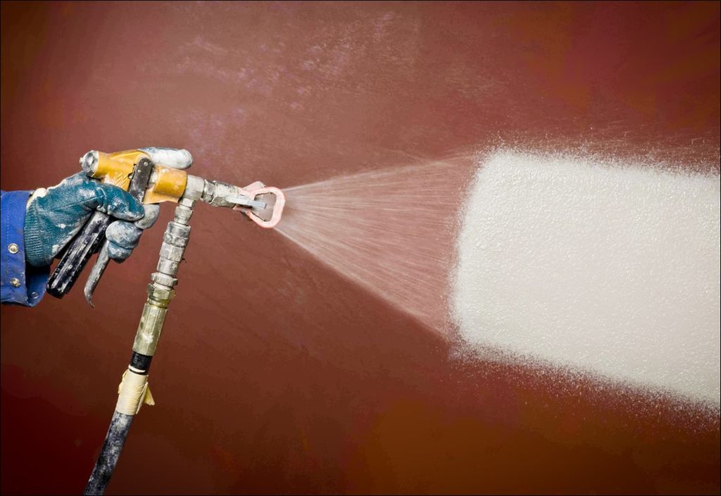 Hånd som holder ei malingssprøyte som påfører hvit maling på en stålkonstruksjon. Foto.