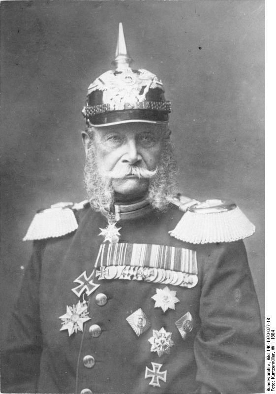 Portrett av den første tyske keisaren, Vilhelm 1. Han har uniform med ei mengd medaljar og utmerkingar, prangande epålettar  og hjelm med ein høg metallpigg på toppen. Foto. 