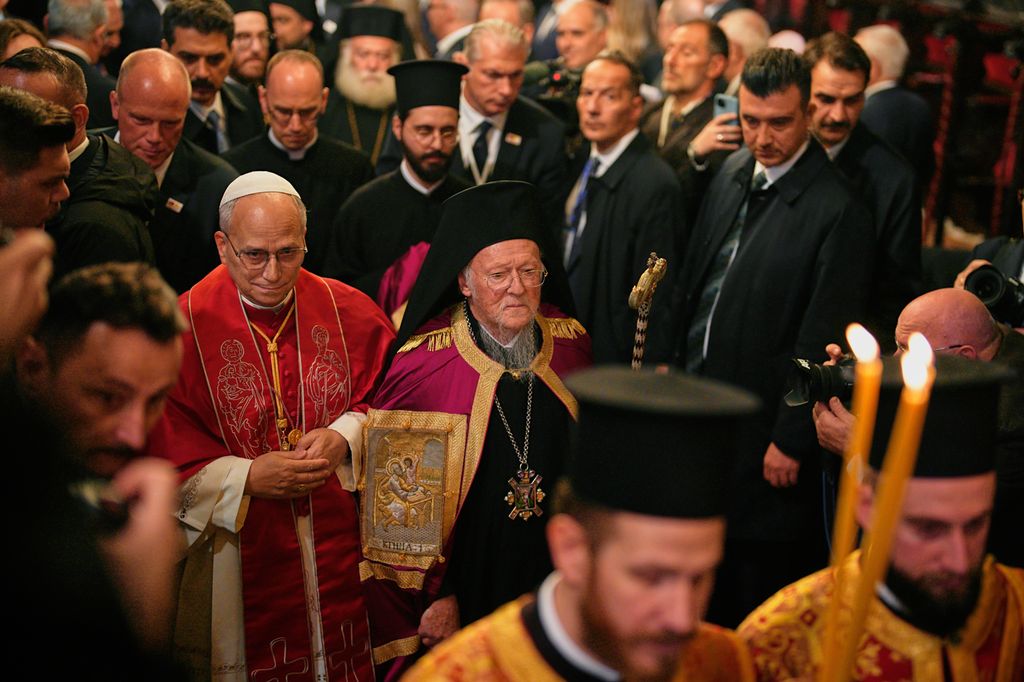 En prosesjon inne i en kirke med religiøse ledere i tradisjonelle liturgiske klær. I forgrunnen er det en person i rød kappe og hvit kalott og en annen i mørkelilla drakt med stort, svart hodeplagg og et ikon rundt halsen. Rundt dem står en større folkemengde. Foto.