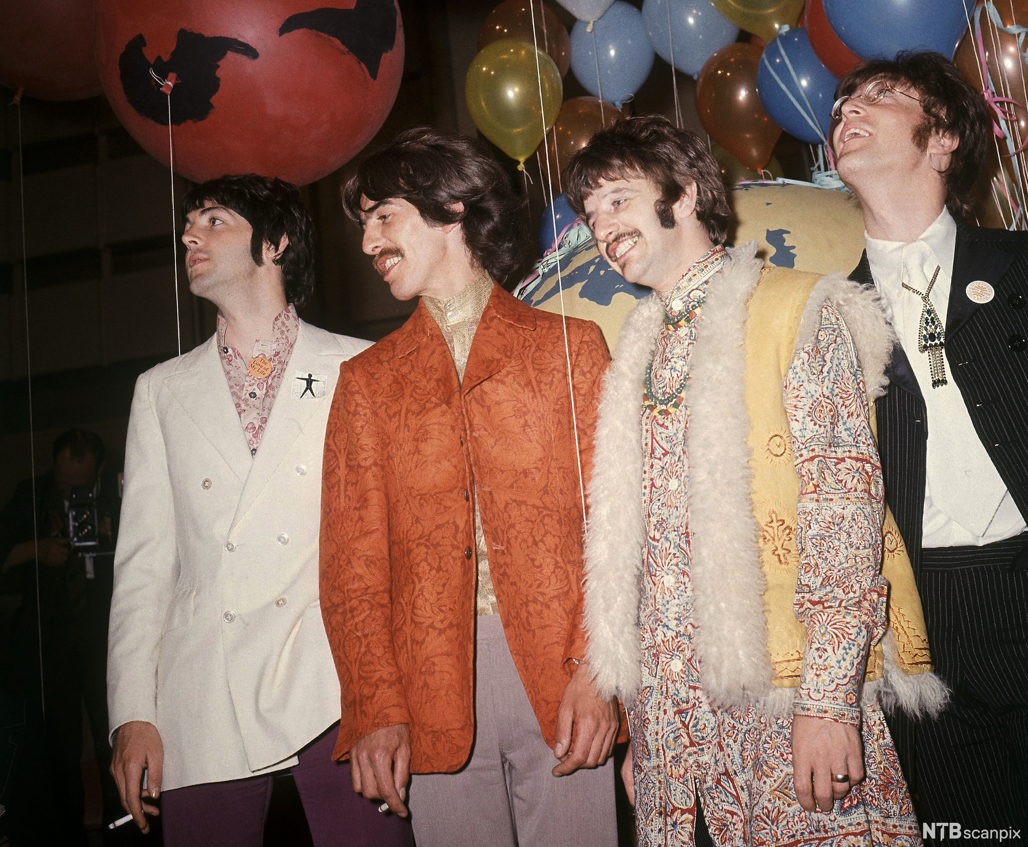 Gruppa The Beatles. Paul McCartney har hvit dobbelspent dress med rosamønstrete skjorte, George Harrison har gullskjorte og en oransje dressjakke med brokademønster, Ringo Starr har skjorte med fargerikt mønster og en vest med hvit pelskant, John Lennon har en klassisk mørk tynnstripete dress og et smykke med stort anheng som henger utenpå slipset. Foto.