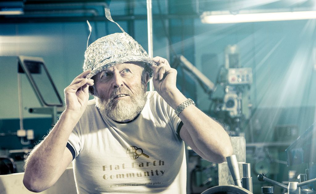 Eldre mann med hatt av aluminiumsfolie og T-skjorte med teksten "Flat Earth Community" på brystet. Foto.