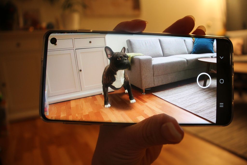 En hånd som holder en mobiltelefon. På mobilskjermen ser vi deler av en stue med en kjøpmannsbenk, en grå sofa med puter og et sofabord. Foto.