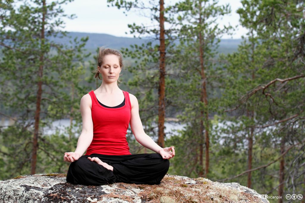 Kvinne med rød topp sitter og mediterer i skogen. Foto.