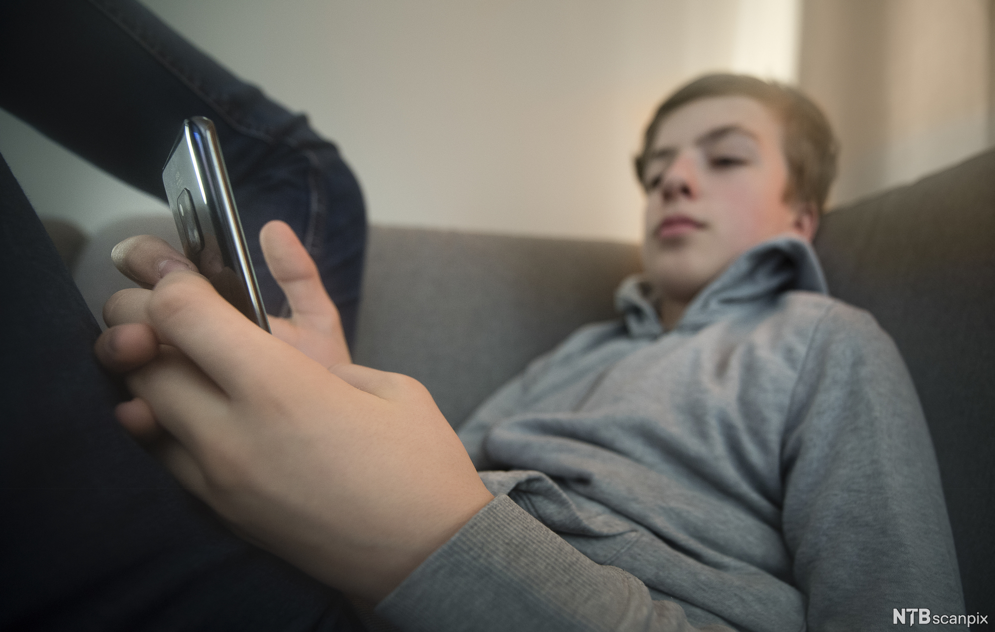 Gutt sitter med beina høyt i en sofa og sjekker mobilen sin. Foto.