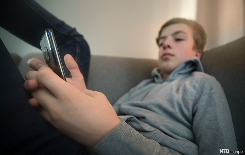 Gutt sitter med beina høyt i en sofa og sjekker mobilen sin. Foto.