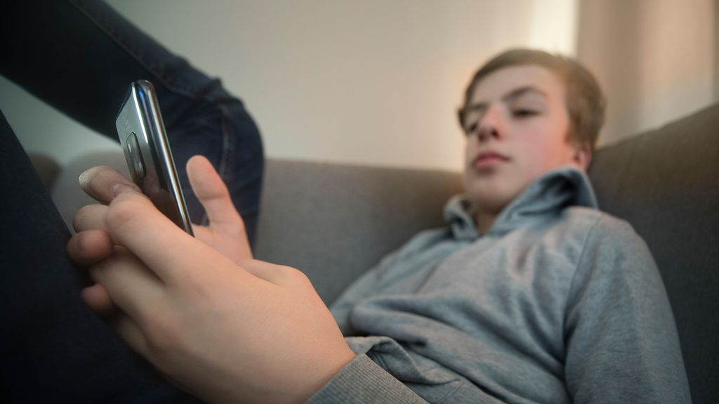 Gutt sitter med beina høyt i en sofa og sjekker mobilen sin. Foto.