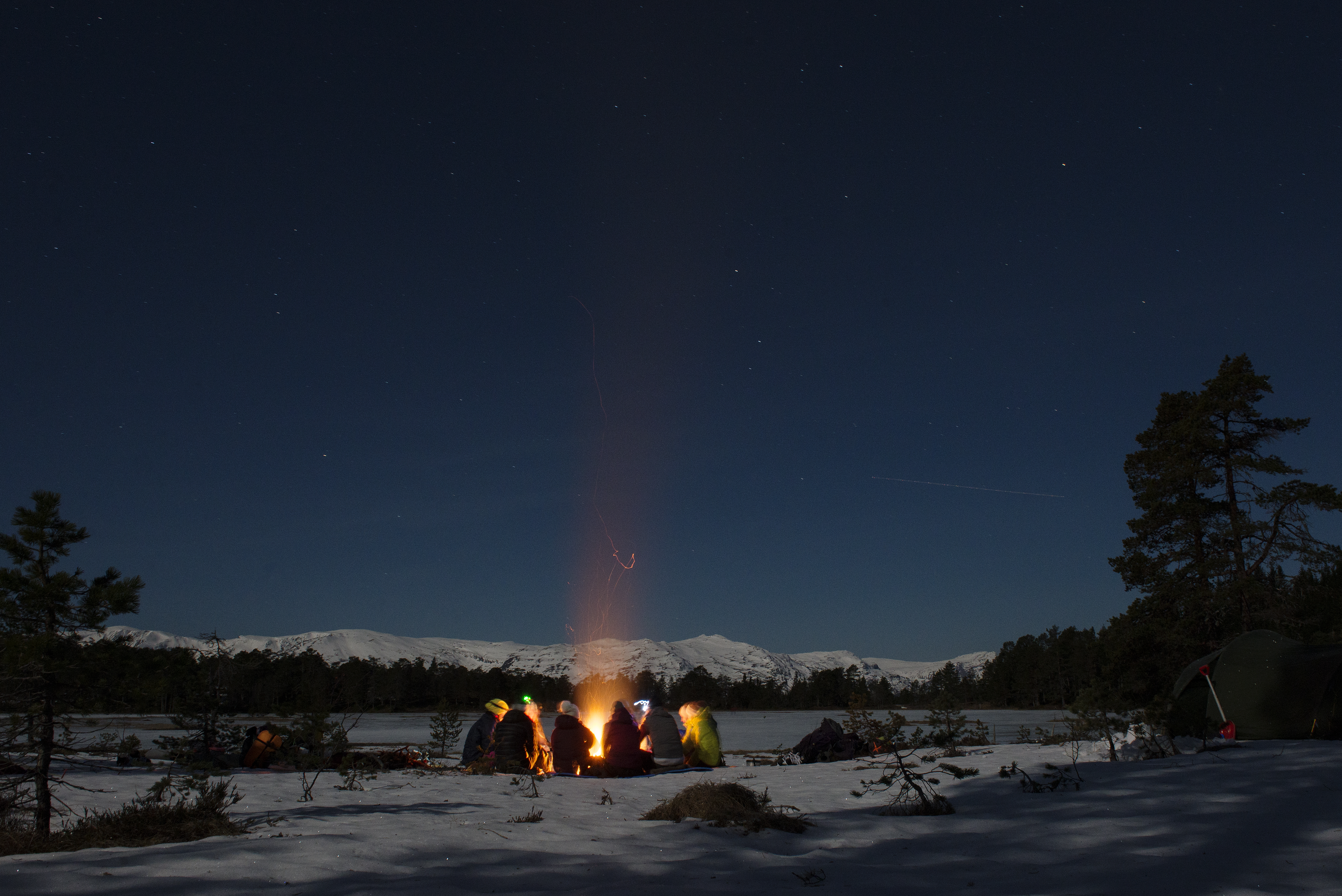 En vinterkveld ute, mørk blå himmel, noen fjelltopper og et islagt vann i bakgrunnen. En gjeng sitter rundt et bål med noen spredte trær rundt seg. Foto. 