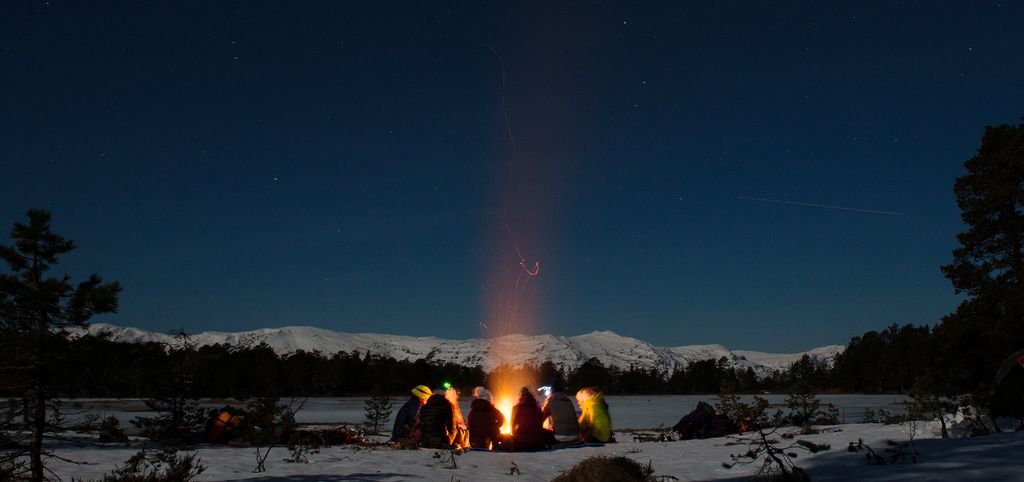 En vinterkveld ute, mørk blå himmel, noen fjelltopper og et islagt vann i bakgrunnen. En gjeng sitter rundt et bål med noen spredte trær rundt seg. Foto.