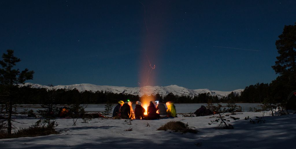 En vinterkveld ute, mørk blå himmel, noen fjelltopper og et islagt vann i bakgrunnen. En gjeng sitter rundt et bål. Foto.