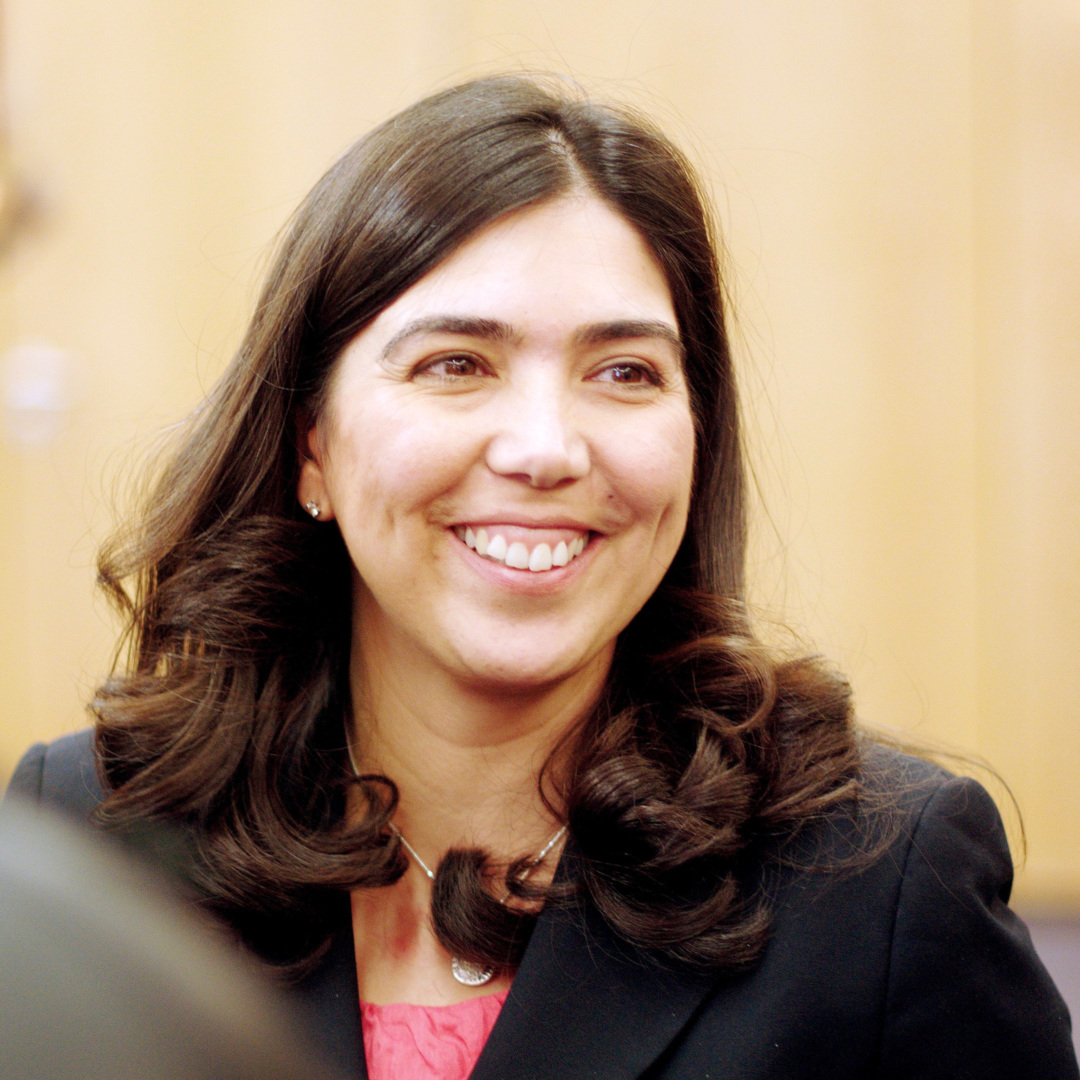 Ei kvinne på rundt 40 år. Ho har langt, svart hår, svarte augebryn og brune auge. Ho har på seg ein antrasittfarga blazer og ein syklamfarga overdel. Fotografi.