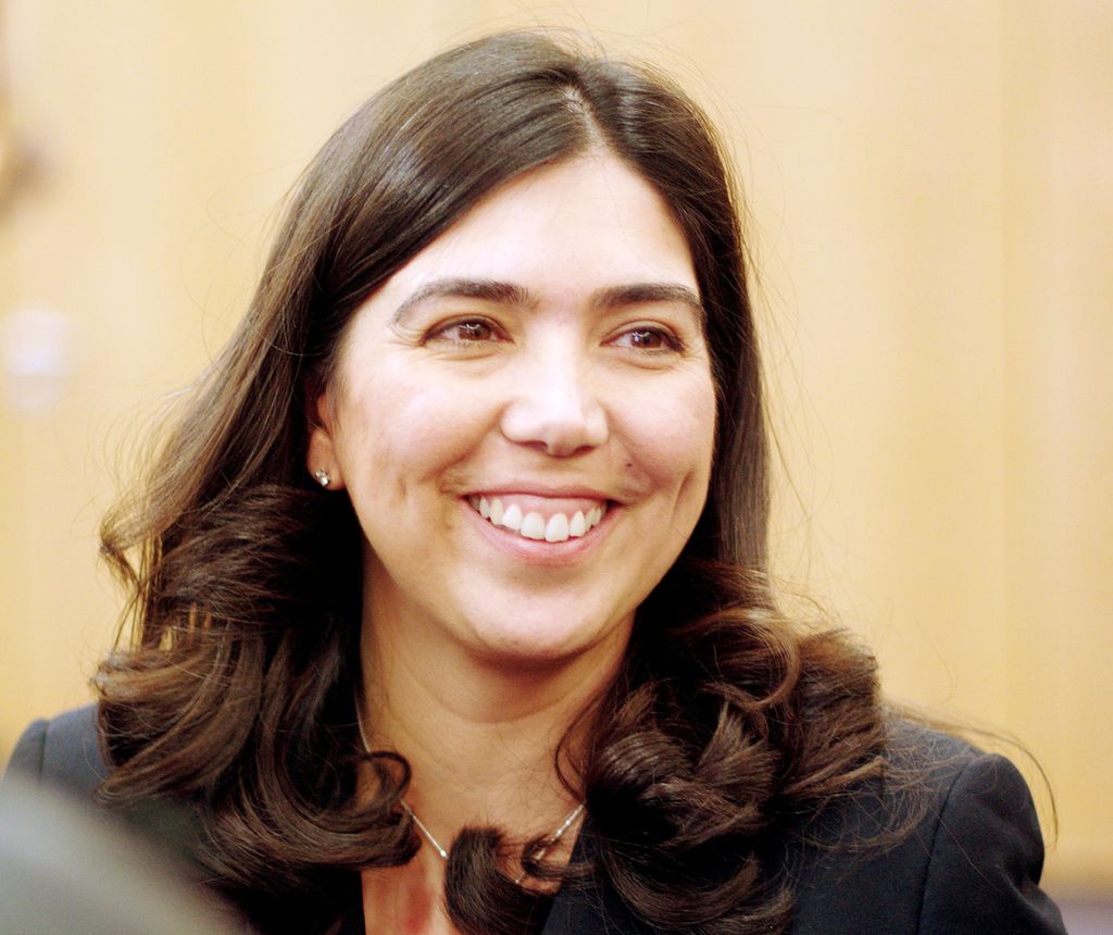 Ei kvinne på rundt 40 år. Ho har langt, svart hår, svarte augebryn og brune auge. Ho har på seg ein antrasittfarga blazer og ein syklamfarga overdel. Fotografi.