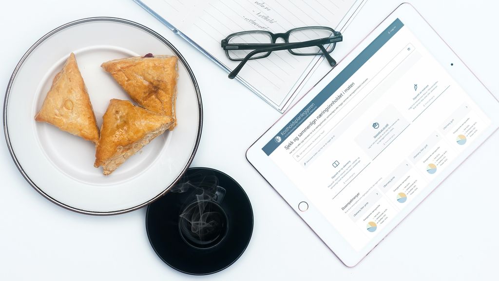 Tallerken med kake, kaffe, notatblokk, briller og et nettbrett med Kostholdsplanleggeren. Foto.