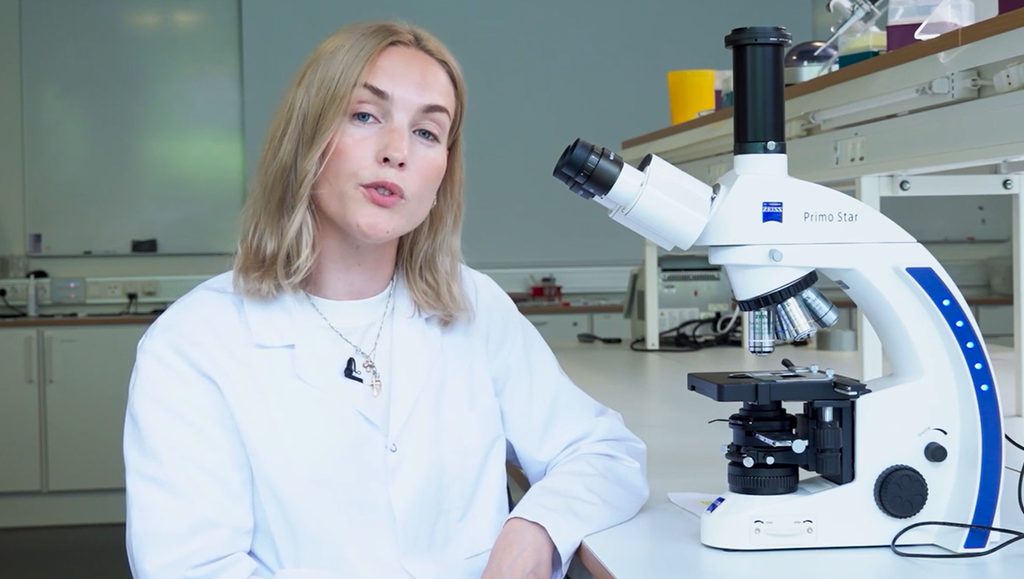 Jente i labfrakk sitter ved siden av et mikroskop. Foto