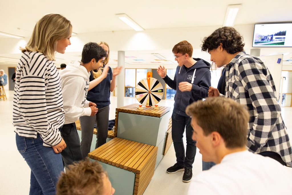 Åtte elever står rundt et lykkehjul i aulaen på en skole. Foto.