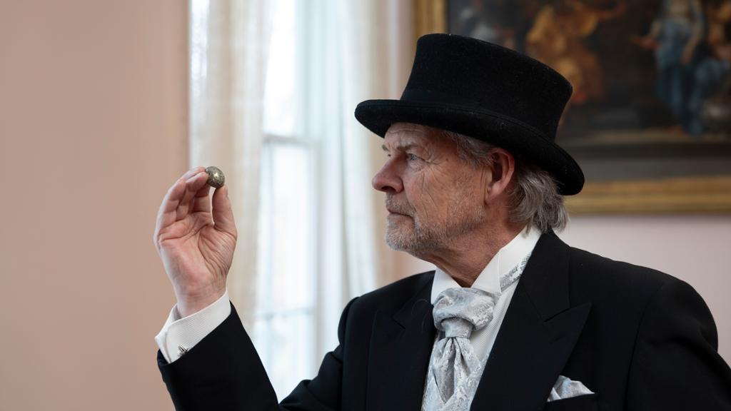 Ein mann kledd i hatt og dress held oppe ein liten stein og ser på han. Foto.