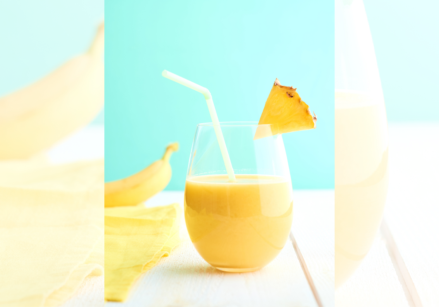 Et sugerør står oppi et glass med gul drikke. På kanten henger det en ananasbit. I bakgrunnen ligger det en banan. Foto.