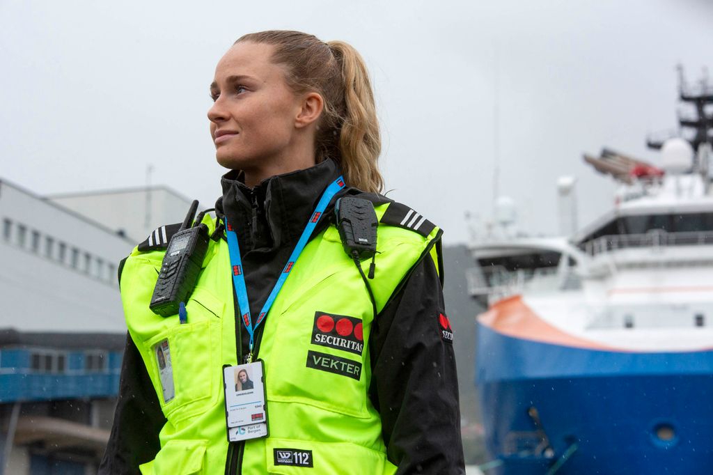 Uniformert kvinnelig vekter med refleksvest og synlig ID-skilt. Ser halvt smilende ut over en kai med flere båter i bakgrunnen. Foto.