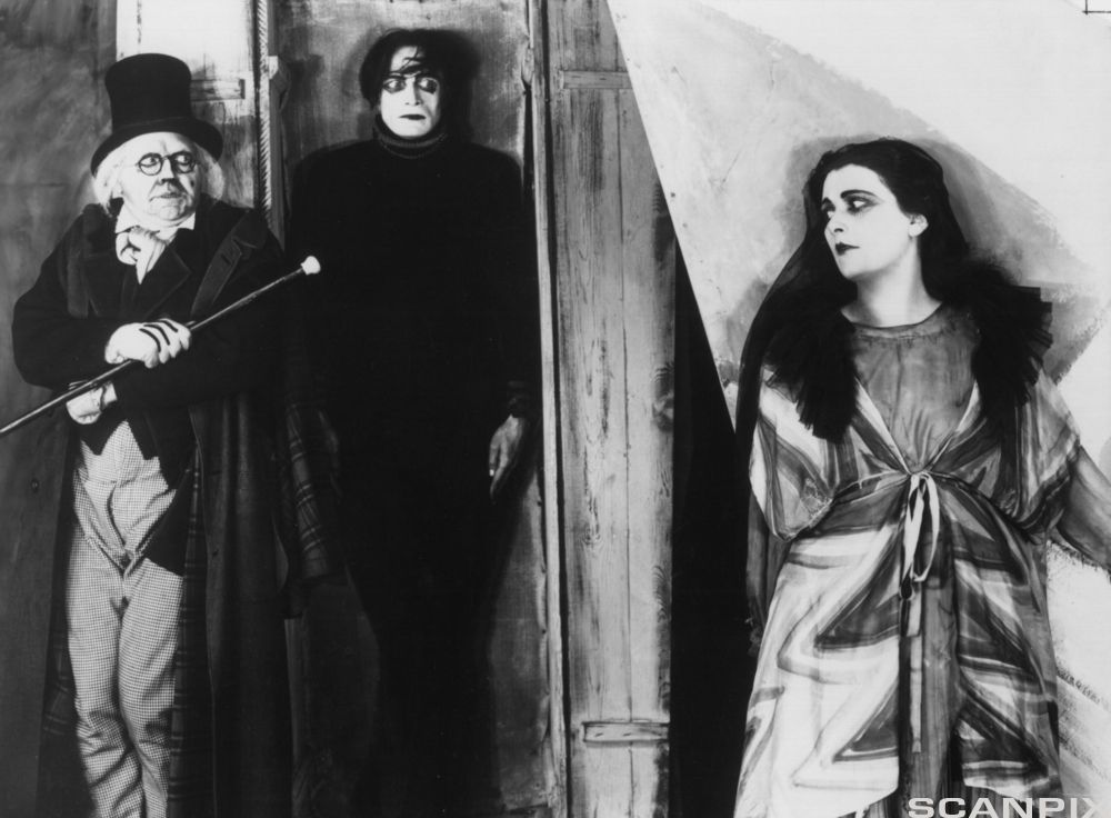 En filmscene viser en mann med flosshatt, briller og dress. Han ser skremt ut og holder en stokk foran overkroppen. Bak han står en helt svartkledd mann med intenst blikk. I forgrunnen lener en svarthåret kvinne seg til en vegg. Bildet har mange og kraftige skygger.