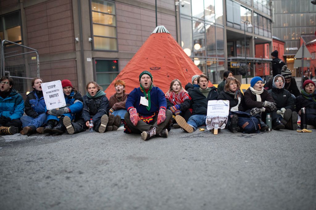 Ungdommar med plakatar sit i ei bygate framfor ein lavvo. Nokon av ungdommane har på seg samedrakt. Foto.