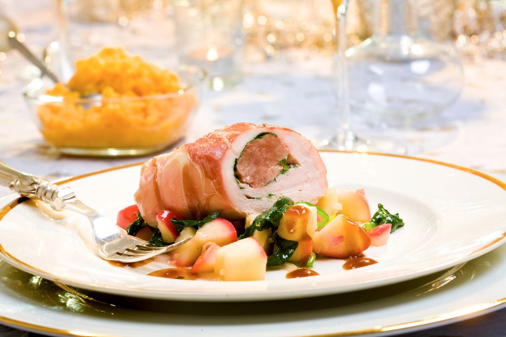 En kyllingfilet kledd i spekeskinke og fylt med farse og urter ligger på en tallerken med eplebiter, spinat og saus. Foto.