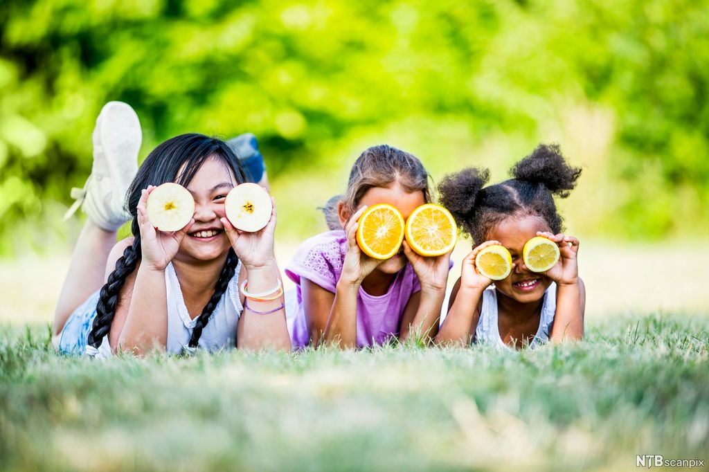 Tre smilende jenter ligger på magen i gresset mens de holder oppskåret frukt foran øynene. Foto.