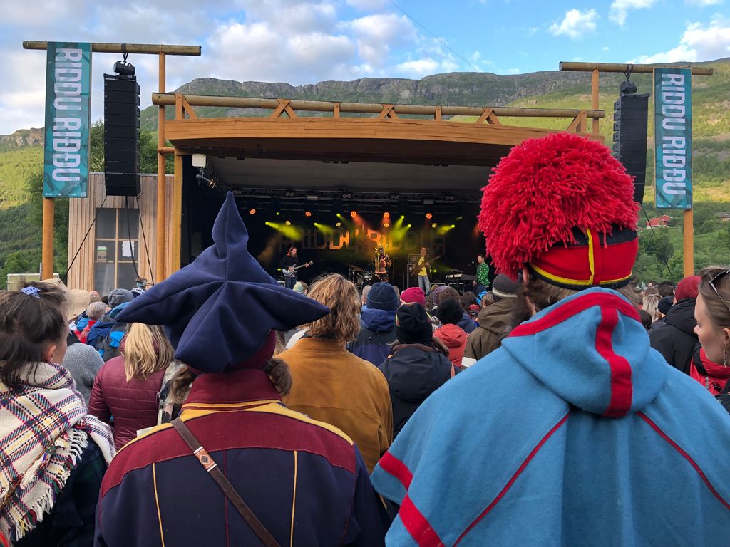 Publikum foran en scene hvor det er konsert. De nærmeste personene har på seg fargerike samiske kofter. Foto.