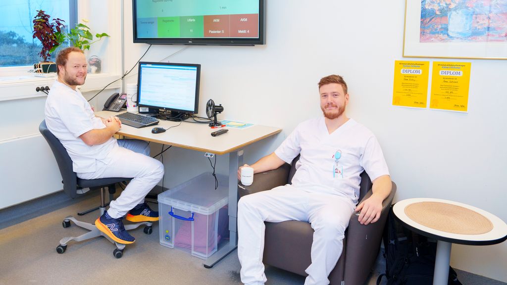 To personer i uniform for helsepersonell smiler mot kamera. Den ene sitter foran en pc, den andre sitter i en stol ved siden av med en kaffekopp i hånda. Foto.