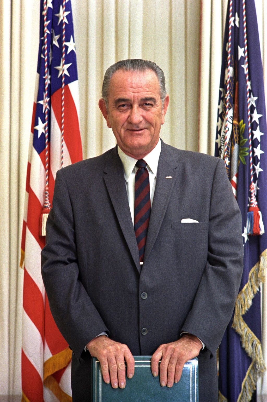 Photo: We see a man with grey hair. He is holding a blue book or folder. Behind him is the US flag and a flag with the seal of the president of the USA.