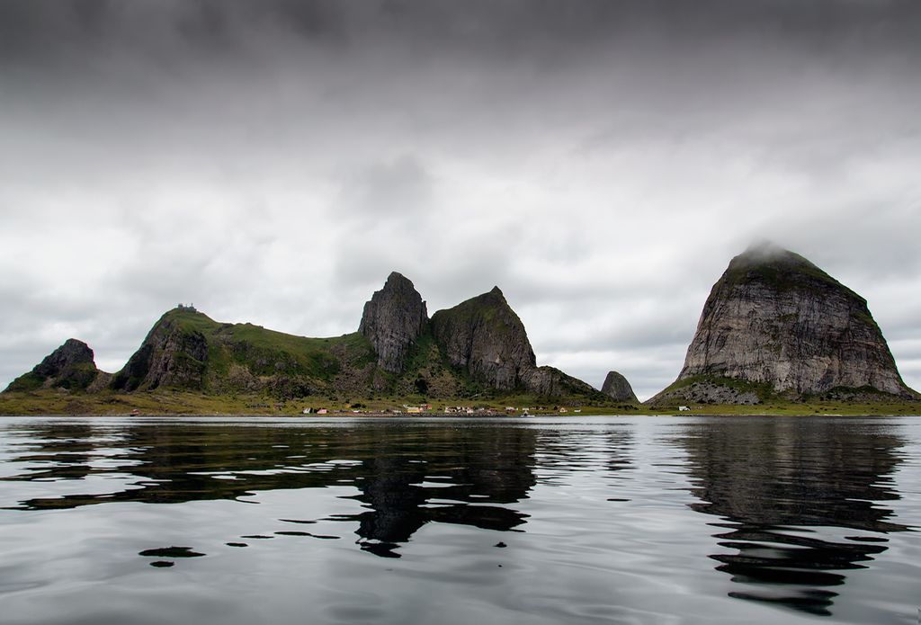 Flere fjelltopper stikker opp av havet. Foran fjellene er det en stripe med flatt landskap med bebyggelse. Bildet viser øya Træna, som ligger utenfor Sandnessjøen. Foto.