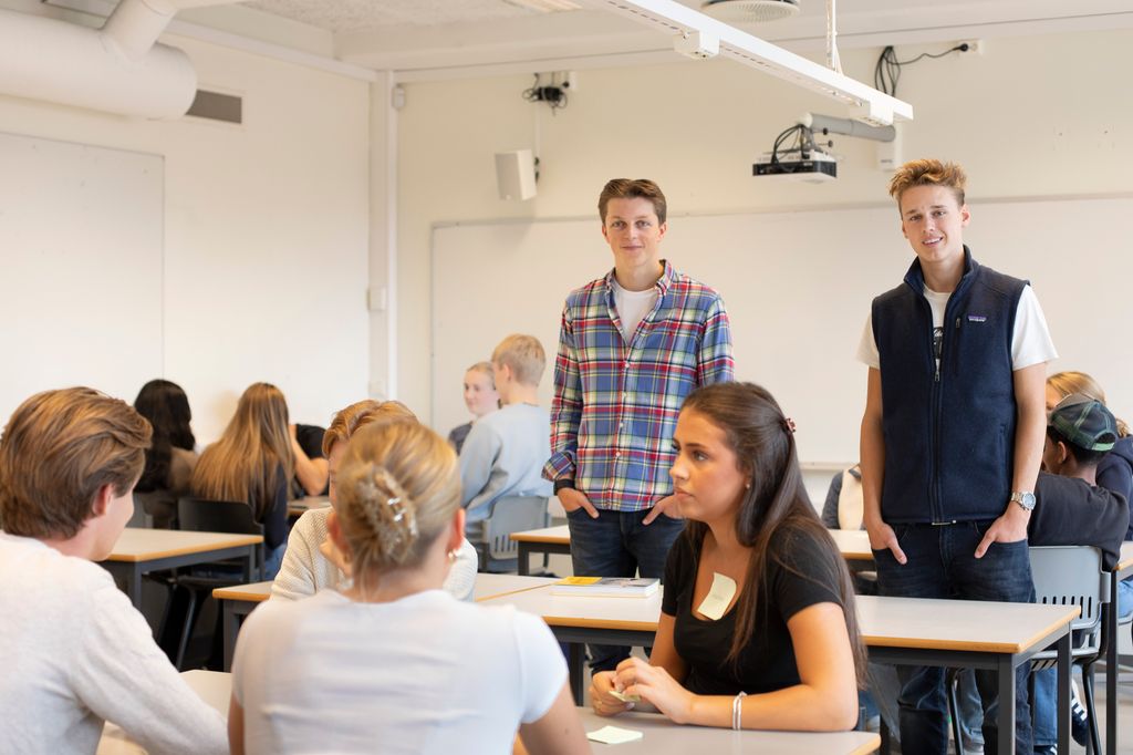 To elever står i klasserommet, mens de andre elevene sitter i grupper. Foto. 