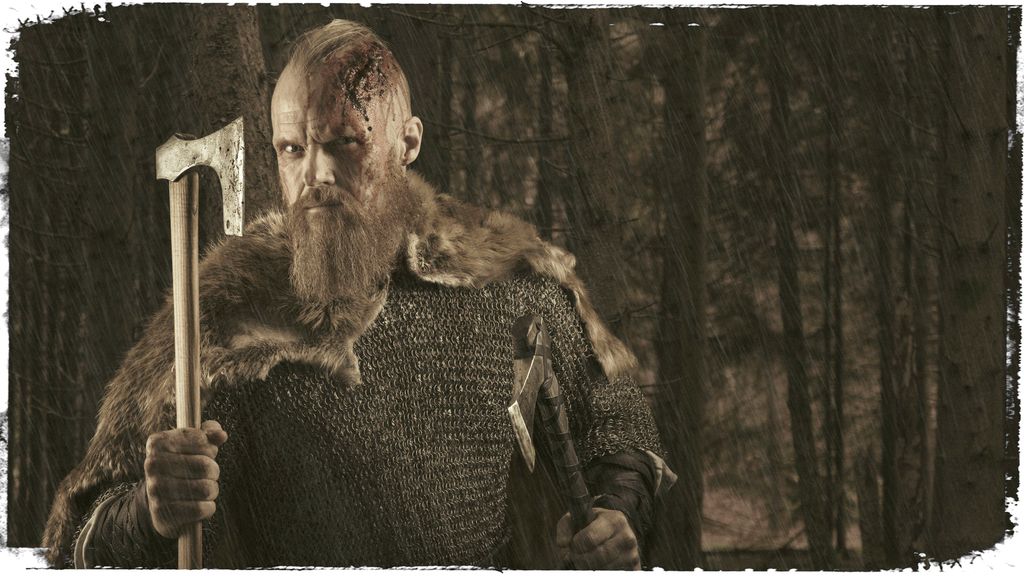 Mann med skjegg, skinnfell rundt halsen og kutt i panna holder ei øks i handa og ser morsk ut. Sort-hvitt-foto.