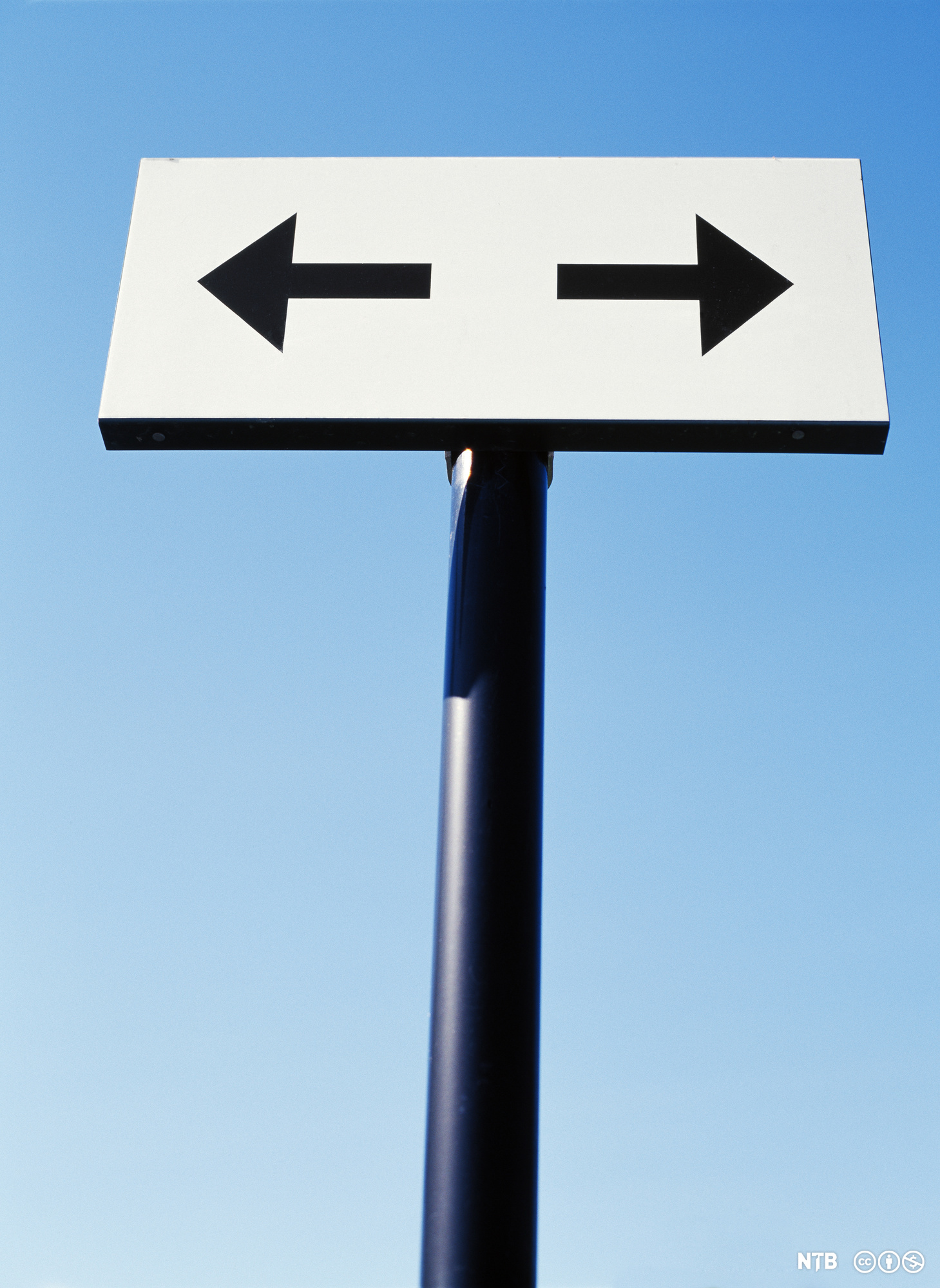 Et skilt viser en svart pil til venstre og en svart pil til høyre på hvit bakgrunn. Bak skiltet er det blå himmel. Foto.