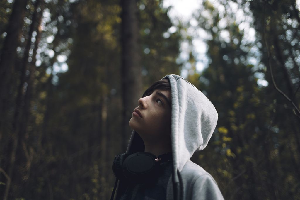 Gutt med hodetelefoner og hettegenser står i en skog og ser opp mot trærne. Foto.