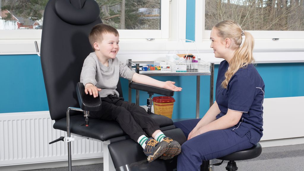 En gutt sitter i en stor pasientstol og smiler til en kvinne som sitter på en krakk foran ham. Hun er kledd i uniform for helsepersonell. Gutten har strukket ut venstre arm og dratt opp genseren. Kvinnen ser på gutten med et vennlig blikk og smil. Foto.