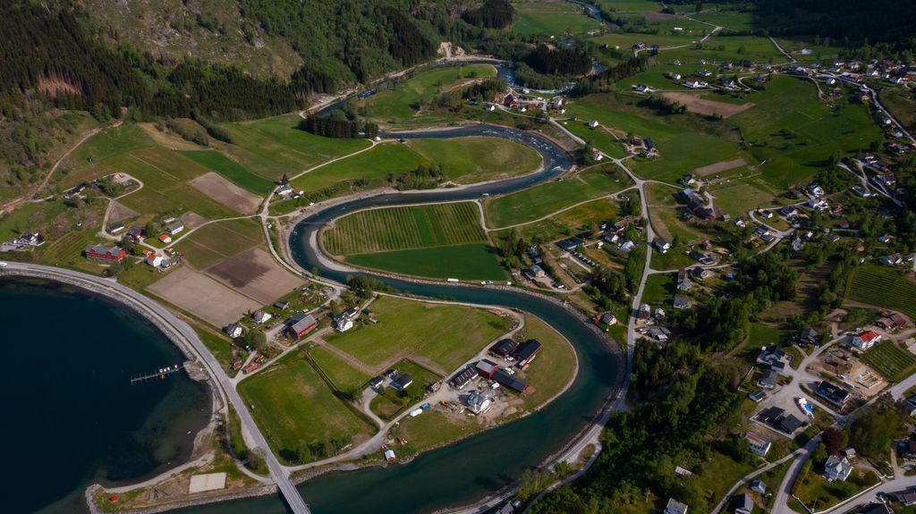 Dronebilete av elv som buktar seg gjennom ein liten tettstad og åkrar ved ein fjord. Foto. 