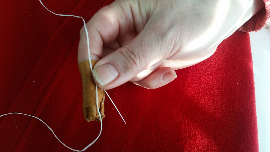 Hand med fingerbjørg av skinn held synål og sytråd. Foto.